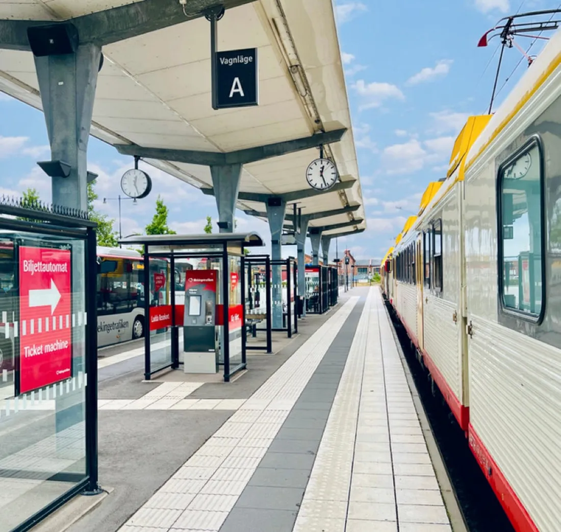Ett stationsläge med väderskydd, biljettautomat och taktilt ledstråk längs plattformen, med ett regionaltåg vid spåret.