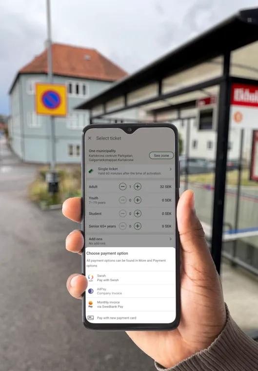 En hand håller en mobiltelefon vid en hållplats. På skärmen väljs biljettyp och betalsätt.